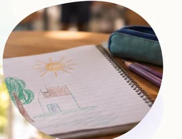 Caderno aberto com um desenho infantil de casa, árvore e sol, ao lado de lápis de cor